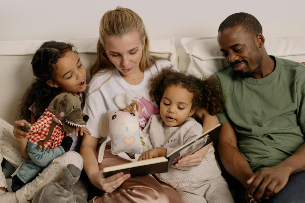 family sitting on a couch with mother reading to two girls as father watches