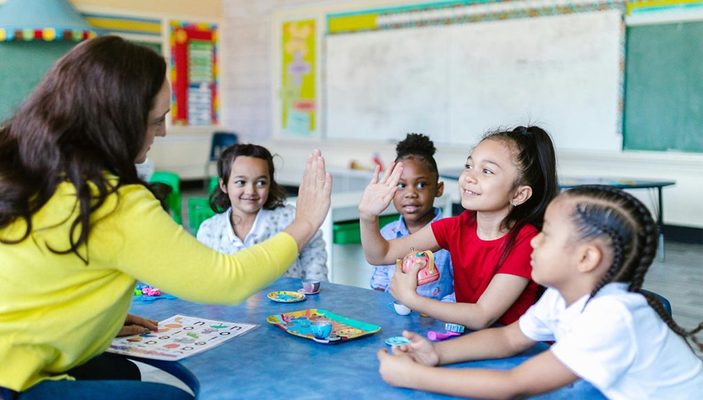 teacher high fiving young students at a table in a classroom