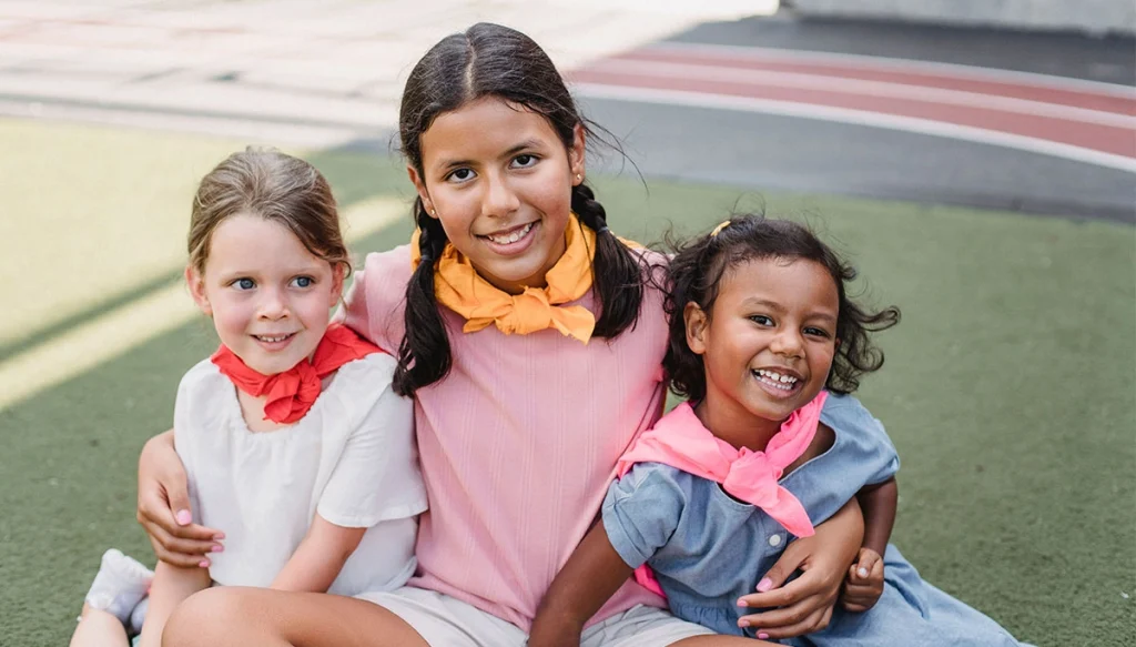 Three diverse girls smiling and sitting on grass, enjoying outdoors.