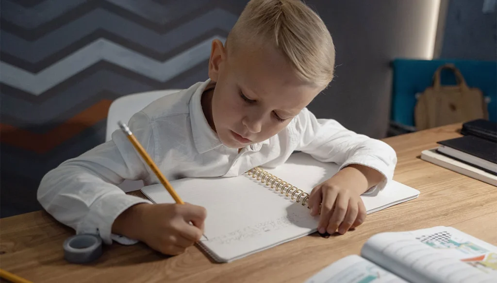 young boy in a white shirt writing in a book on a lamp lit table